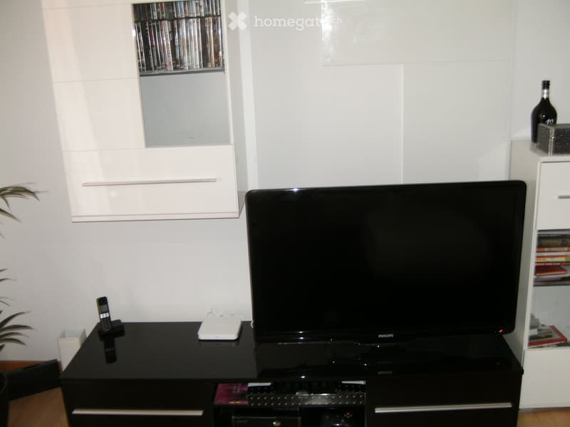 Modern living room with a large flat-screen TV on a black stand, a black table with a cellphone and a white device, and built-in white cabinets with books and DVDs.