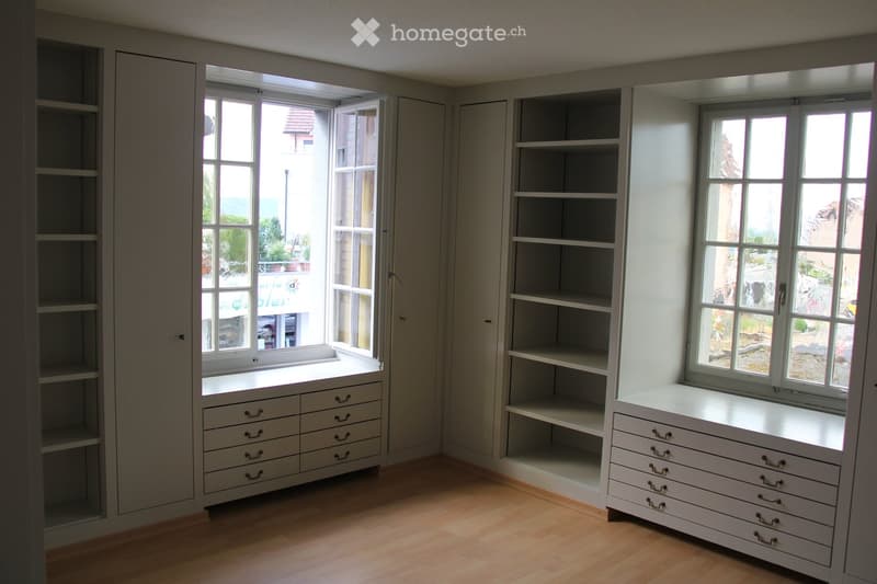 Empty room with built-in white cabinets and shelves, large windows with open blinds, and a wooden floor.