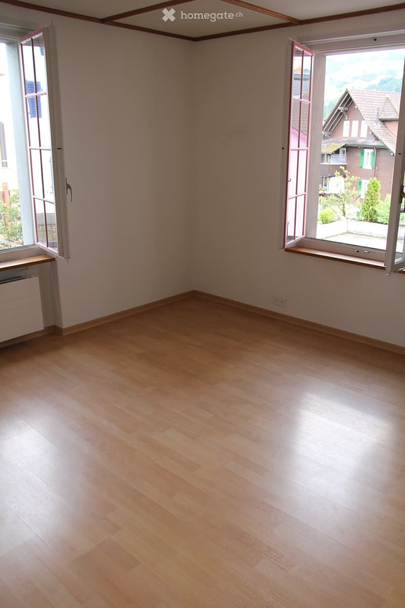 Empty room with light wooden flooring, white walls, and large windows with open shutters, offering a view of the outside area.