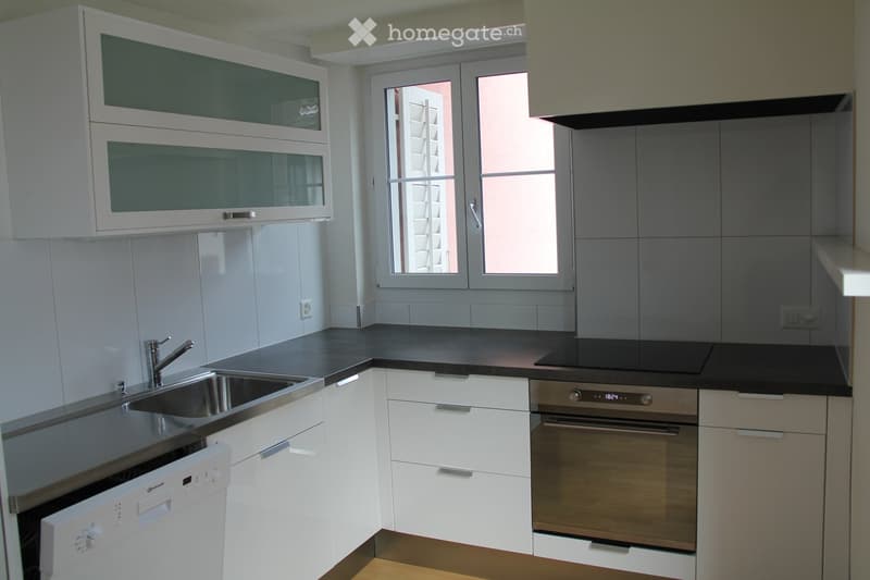 Modern kitchen with white cabinets, black countertop, stainless steel sink, dishwasher, and oven. Features a large window with blinds.