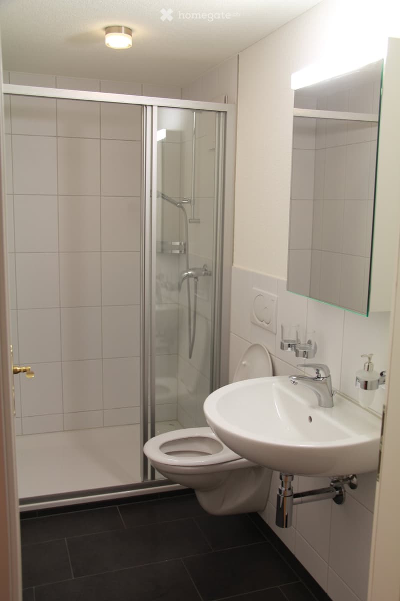 Modern bathroom with white tiles, a toilet, a sink with a modern faucet, a shower with a glass enclosure, and a mirror with built-in lighting.