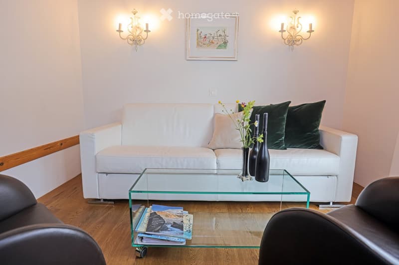 Modern living room with a white sectional sofa, green throw pillows, a glass coffee table, and two black armchairs. The room features wooden flooring, wall-mounted candle holders, and a framed picture on the wall.