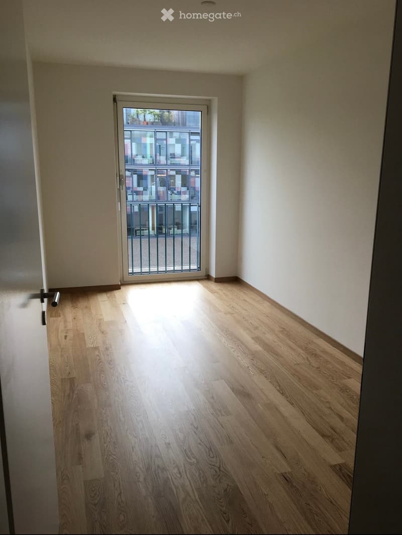 Empty room with light wooden flooring and white walls, featuring a sliding glass door leading to a balcony with a metal railing.