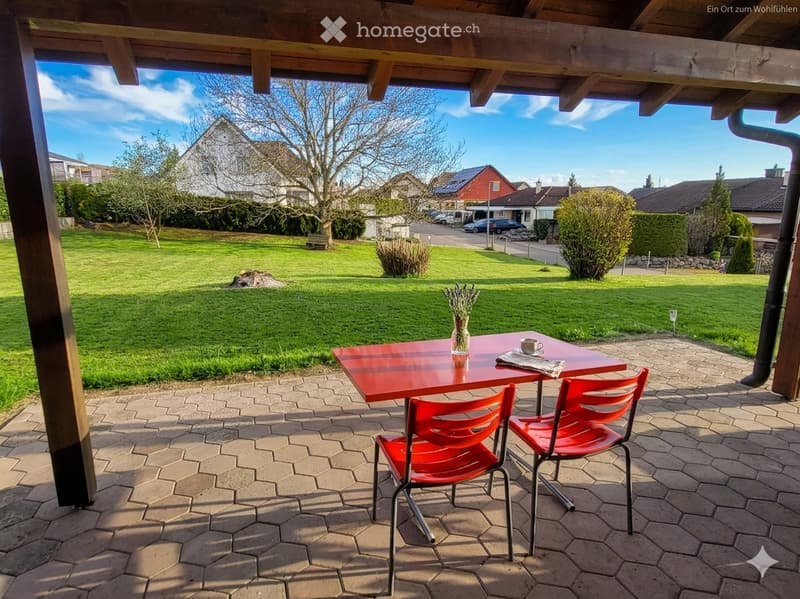 A covered outdoor seating area with a red table and two red chairs, overlooking a well-maintained lawn and residential neighborhood.