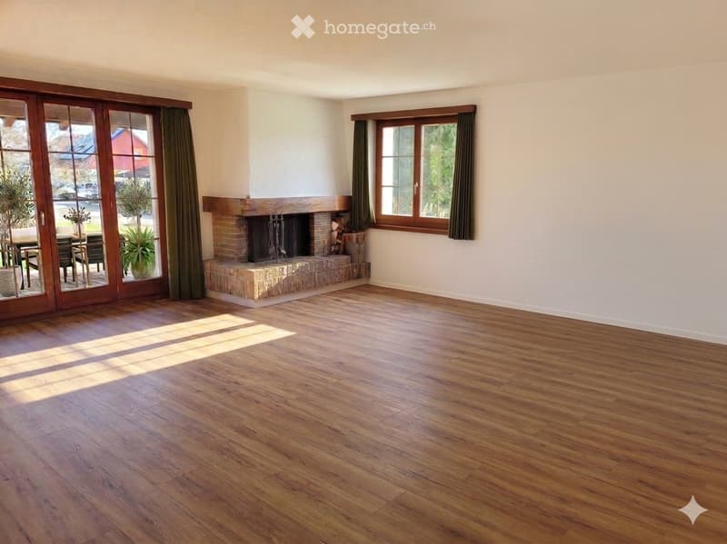 Empty living room with wooden flooring, fireplace, large windows, and sliding glass doors leading to an outdoor area.