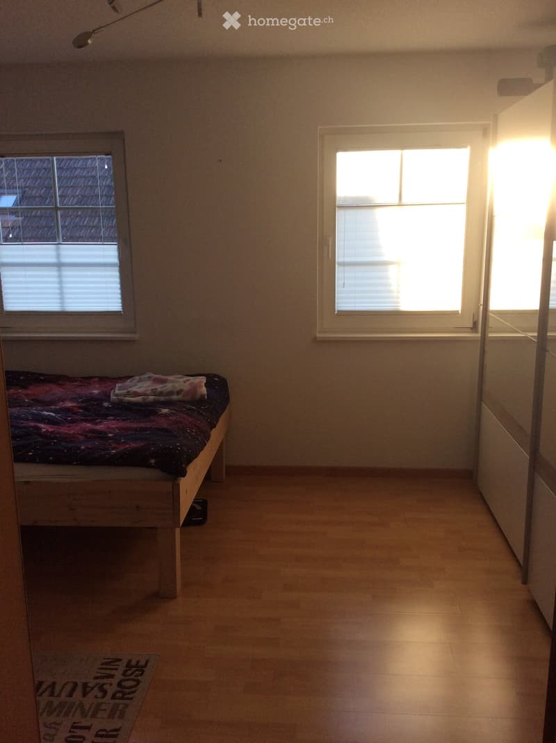A small, furnished bedroom with a wooden bed, a colorful blanket, a rug, and large windows allowing natural light to enter.