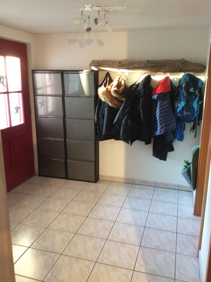 Empty hallway with tiled floor, coat rack with jackets and bags, and a mirrored cabinet on the left side.