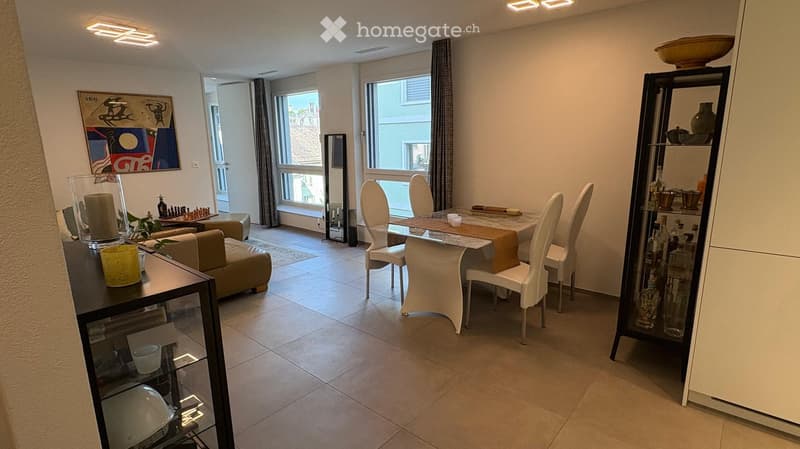 A modern, well-lit living and dining area with a sofa, dining table, chairs, shelves, and decorative items.