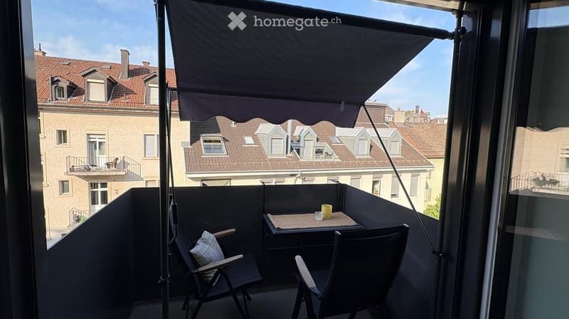 Balcony with a small table, two chairs, and a cup, featuring a retractable awning and a view of neighboring buildings.