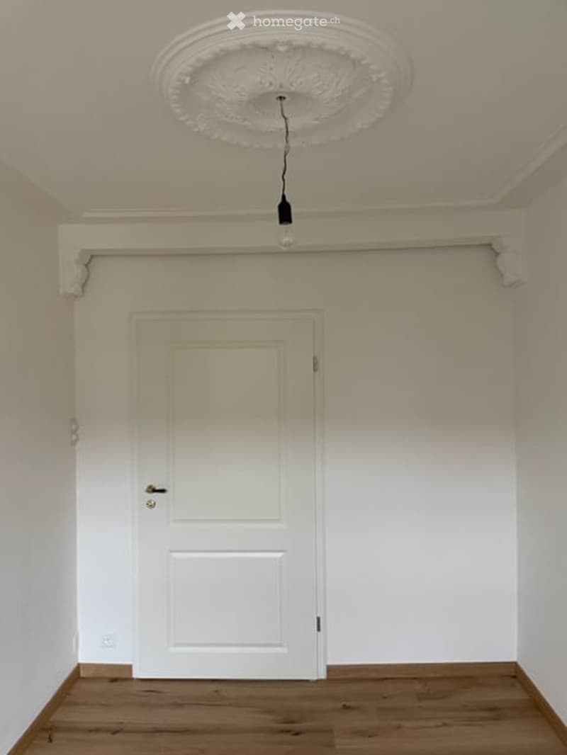 Empty room with white walls, wooden floor, white door, and a decorative ceiling light fixture.