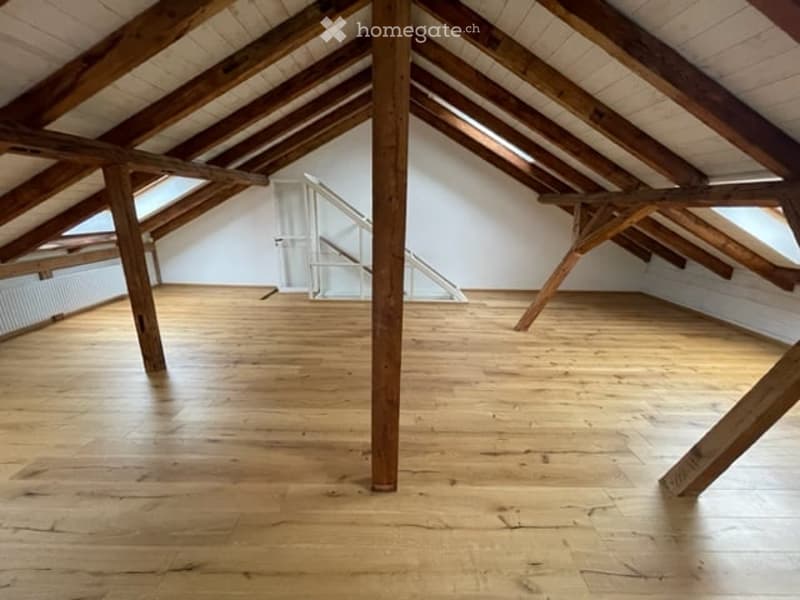 Empty attic space with wooden beams and polished wooden floor, featuring exposed wooden beams and a small staircase leading up.