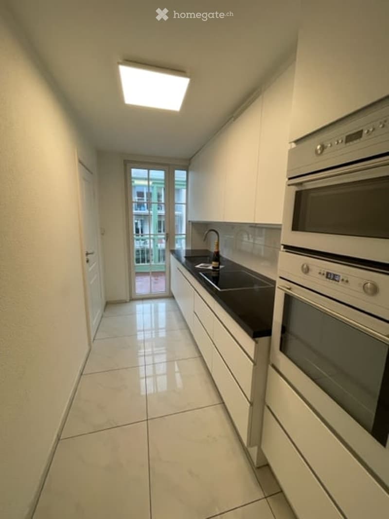 Modern kitchen with white cabinets, black countertops, a sink, and built-in oven. Large window offers a view of the balcony and outdoor area.