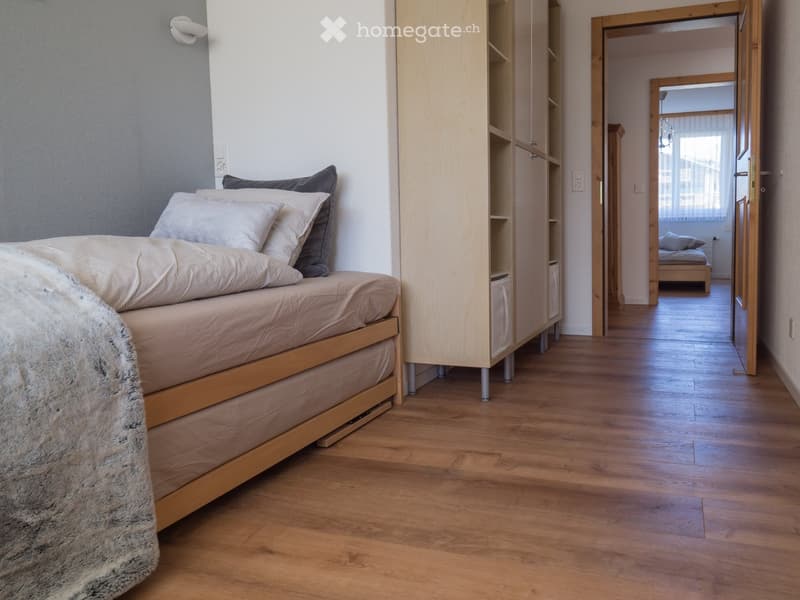 Cozy bedroom with a wooden bed, pillows, a fur throw, and a light wooden wardrobe. The room has wooden flooring and an open doorway leading to another room.