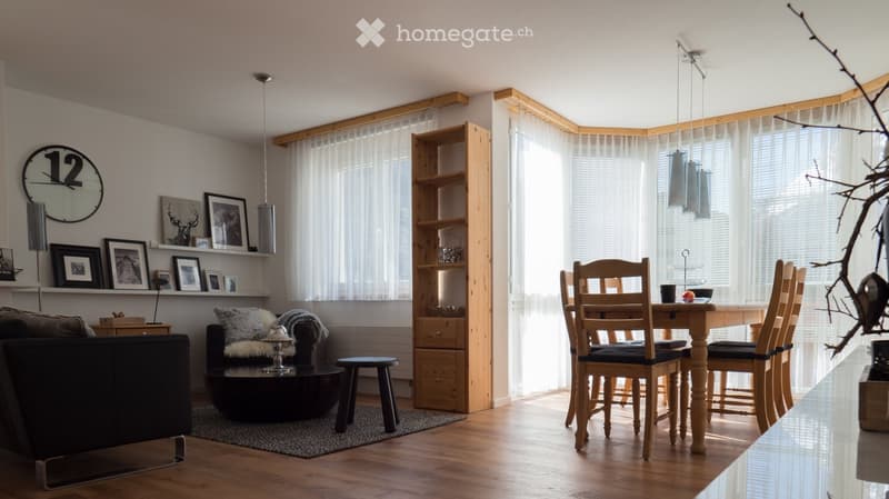 Bright living and dining area with wooden flooring, large windows with white curtains, a black sofa, a round coffee table, a dining table with four chairs, a wooden shelf, and a clock on the wall.