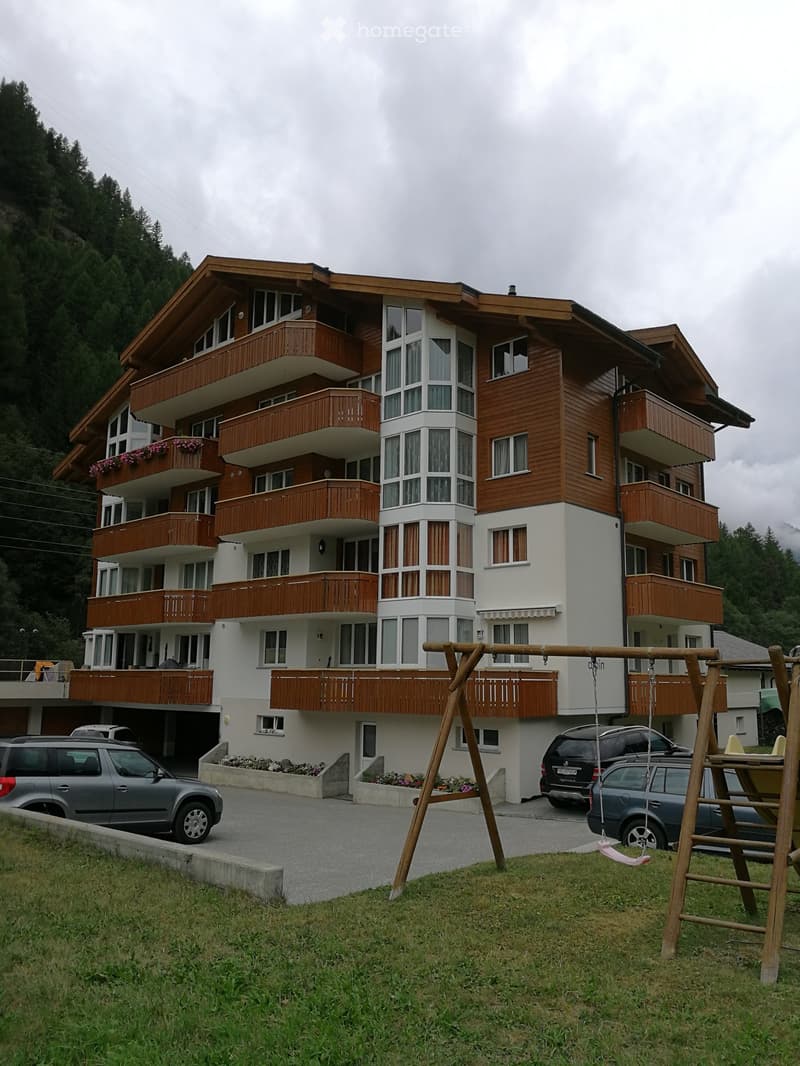 A multi-story residential building with wooden balconies and large windows, surrounded by a parking area and a small playground with a swing.
