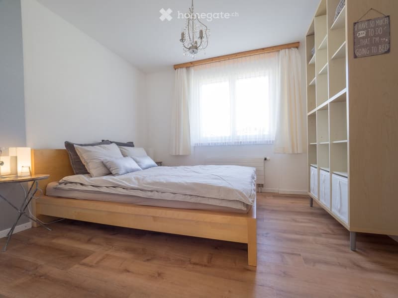 Cozy bedroom with wooden flooring, a neatly made bed with pillows and blankets, a nightstand with a lamp, a small table with a lamp, and a built-in bookshelf with storage compartments.
