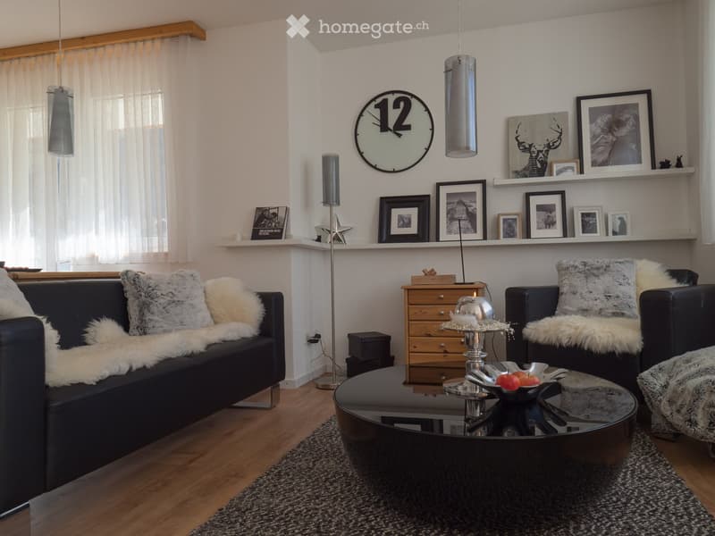 Modern living room with dark leather sofa, armchair, glass coffee table, floor lamp, clock, and decorative shelves with picture frames.
