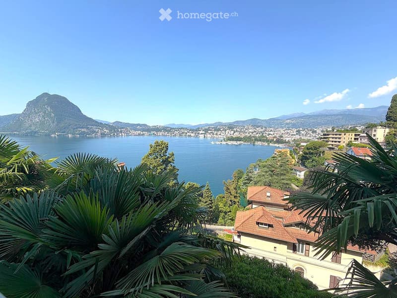 A scenic view of a lake surrounded by mountains, with palm trees in the foreground and a house visible on the right side.
