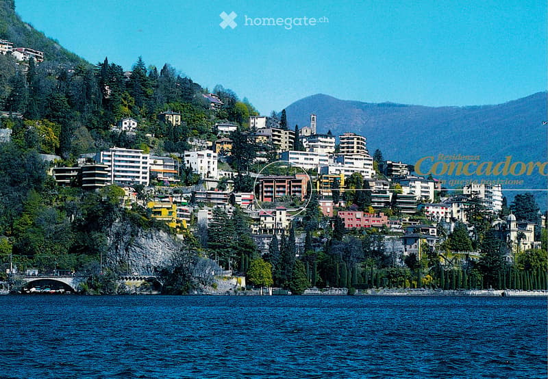 A scenic view of a town built on a hillside overlooking a lake, with numerous buildings and lush greenery.