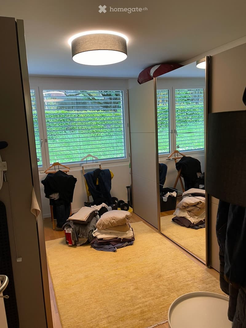 Small, well-lit room with a large mirror, clothing rack, and neatly stacked bedding on the floor.