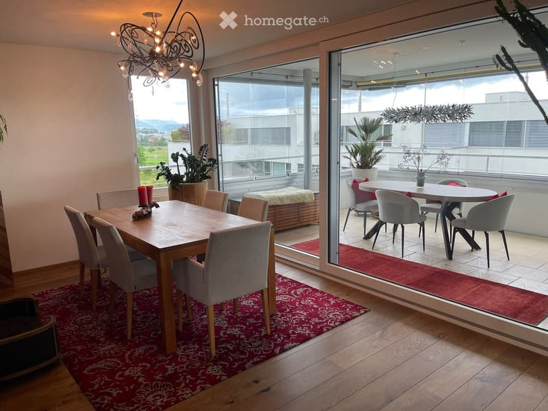 Bright dining area with wooden dining table, six chairs, red patterned rug, chandelier, and large windows offering a scenic view. Adjacent balcony features a round table with four chairs, red cushions, and potted plants.