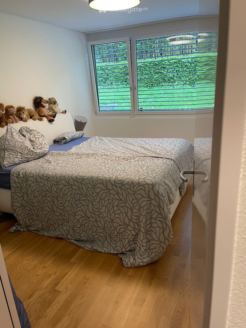 Cozy bedroom with a double bed, patterned bedspread, pillows, a small sofa, and stuffed animals on the headboard. Large window with blinds offers a view of greenery.
