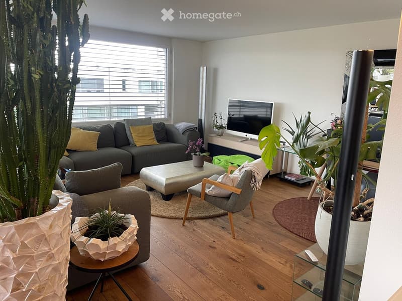 Modern living room with wooden flooring, large window, grey sofa, coffee table, armchairs, ottoman, TV, and various plants.