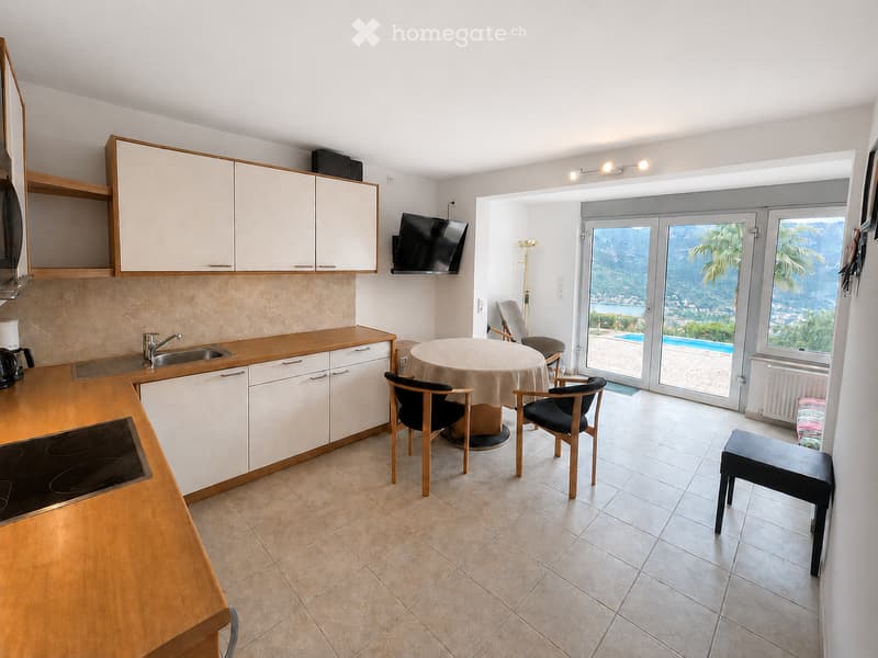 A modern kitchen and dining area with white cabinets, a wooden countertop, an electric stove, a sink, and a coffee maker. A round dining table with chairs is placed near a large sliding glass door that opens to a view of a pool and outdoor area.