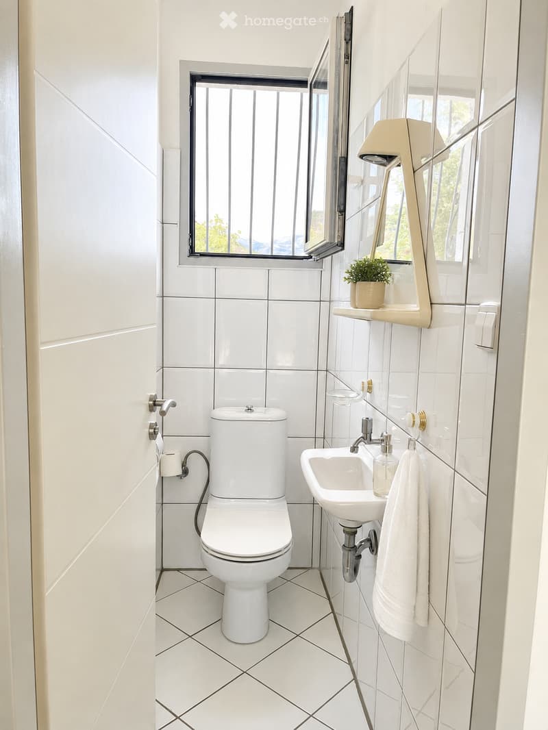 Modern bathroom with white tiles, a toilet, a sink with a mirror, a towel, and a small plant on the windowsill.