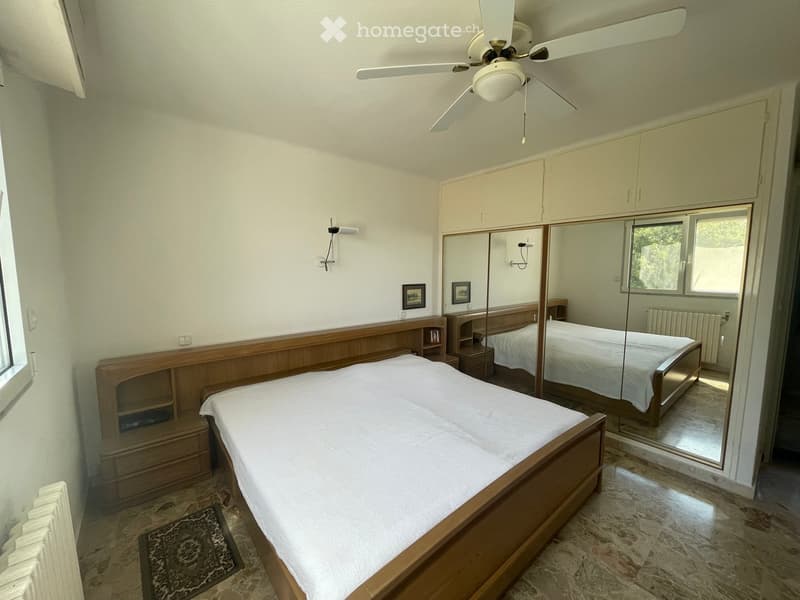 Well-lit bedroom with a wooden bed, mirrored wardrobe, nightstand, and ceiling fan.