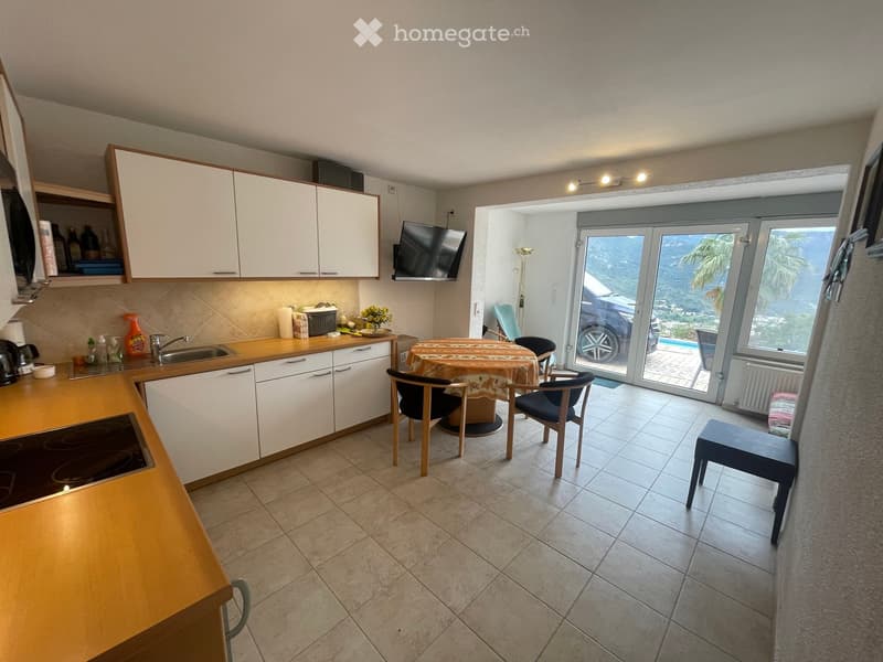 Bright kitchen with white cabinets, wooden countertops, induction stove, sink, and a small dining area with a table and chairs. A television is mounted on the wall, and a large window offers a scenic view of the sea and surrounding landscape.