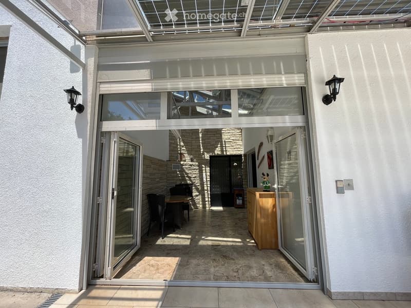 A bright, modern entryway with large glass doors leading to an indoor seating area. Inside, there is a wooden table with chairs, a wooden cabinet, and a small decorative statue on top.
