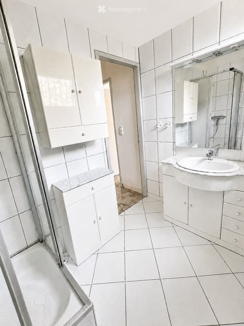 Modern bathroom with white tiled walls and floor, white cabinets, a sink with a mirror, and a glass-enclosed shower area.