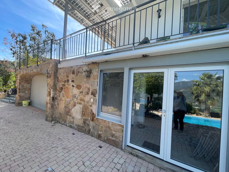 Exterior view of a modern two-story house with stone walls, large glass doors, and a balcony with metal railings. A swimming pool is visible through the glass doors, and the surrounding area includes a paved brick walkway and greenery.