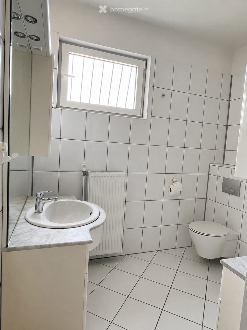 Modern bathroom with white tiled walls and floor, a white sink with a chrome faucet, a toilet, a radiator, and a window with frosted glass panes.