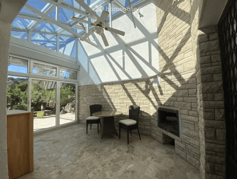 Bright sunroom with a stone fireplace, dining table, chairs, ceiling fan, and large windows overlooking a scenic outdoor area.