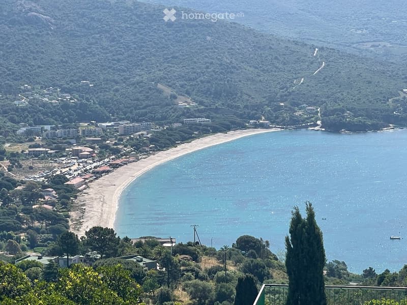 A scenic coastal view showing a sandy beach, calm sea, and a town with buildings nestled among trees and hills.