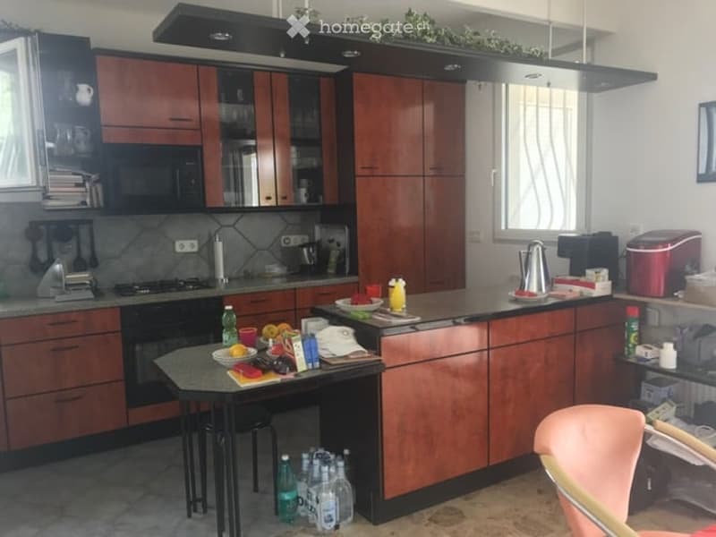 A modern kitchen with wooden cabinets, stainless steel appliances, and a central island with a table and chairs. The counter is cluttered with various items including bottles, a coffee maker, and food items.