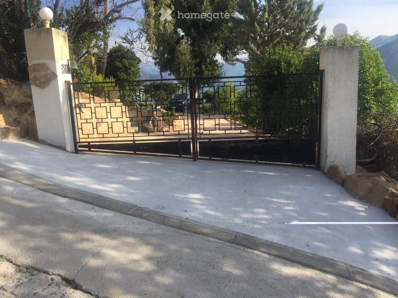 Entrance gate with decorative metalwork, flanked by stone pillars and greenery, leading to a driveway.