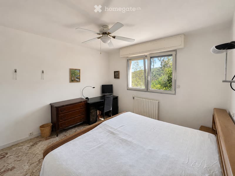 Cozy bedroom with a wooden bed, desk with a computer, a dresser, and a wicker basket on the floor. Large windows offer a view of greenery outside.