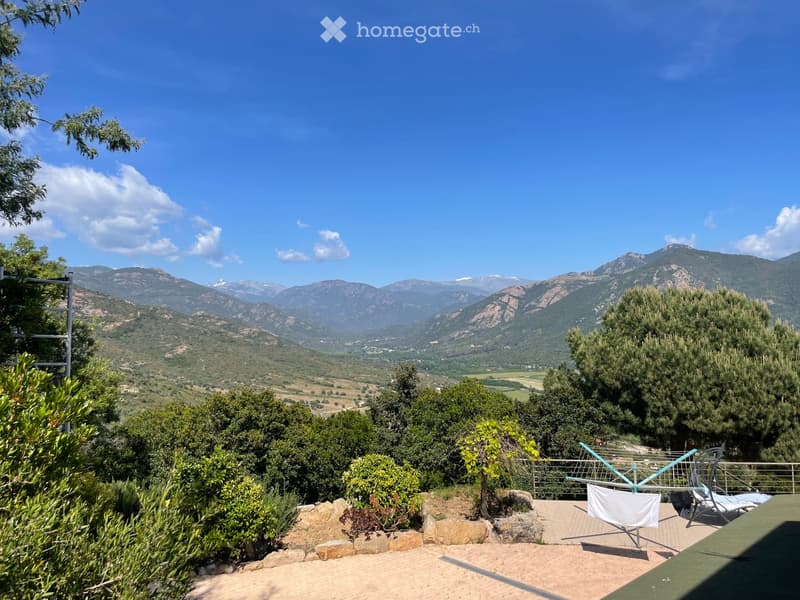 A scenic outdoor area with a panoramic mountain view, surrounded by lush greenery and trees, featuring a patio with lounge chairs and a small table.
