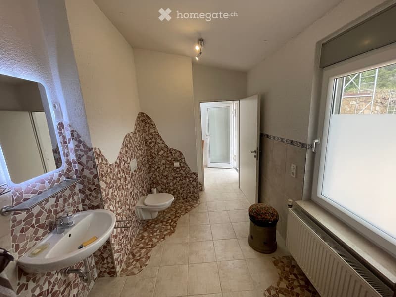Bathroom with mosaic tile accents, a sink, toilet, radiator, and a trash can. The room has large windows and a door leading to another space.