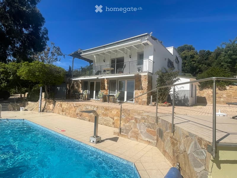 Modern two-story house with a swimming pool, stone terraces, outdoor seating, and a balcony on the upper level.