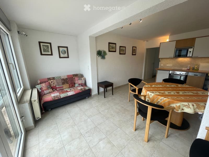 A cozy, well-lit living and dining area with a colorful patchwork sofa, a round dining table with chairs, a small black stool, and a compact kitchen area featuring a microwave, oven, and dishwasher. The space is decorated with framed pictures and a small