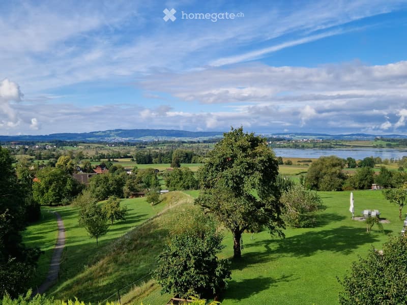 A panoramic view of a lush green garden with trees, a pathway, and a scenic landscape featuring fields, a lake, and distant hills under a partly cloudy sky.