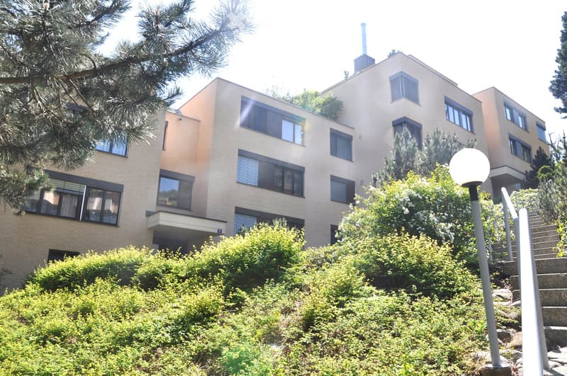 A multi-story residential building with a modern design, featuring large windows, a balcony, and surrounded by greenery and bushes.