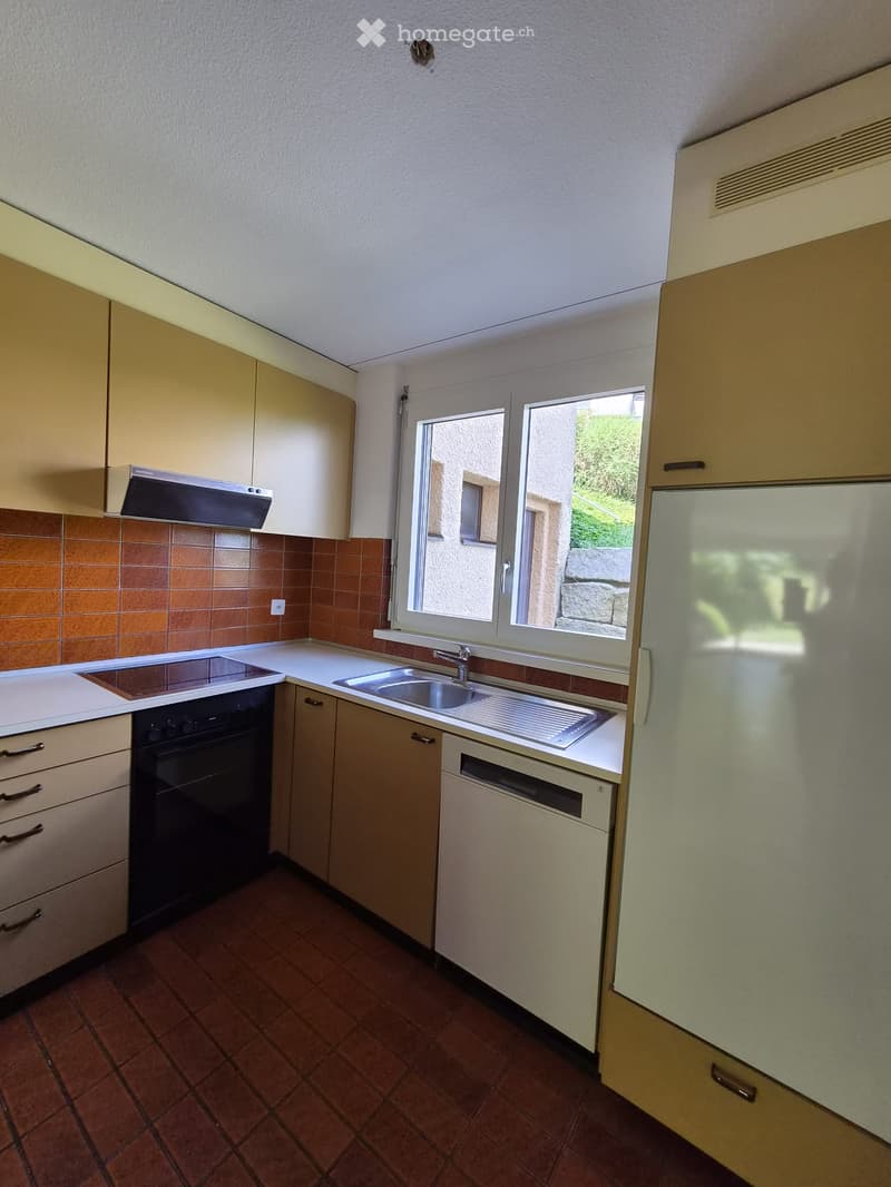 A compact kitchen with light-colored cabinets, a white countertop, and a window overlooking a garden. Features include a refrigerator, oven, sink, dishwasher, and range hood.