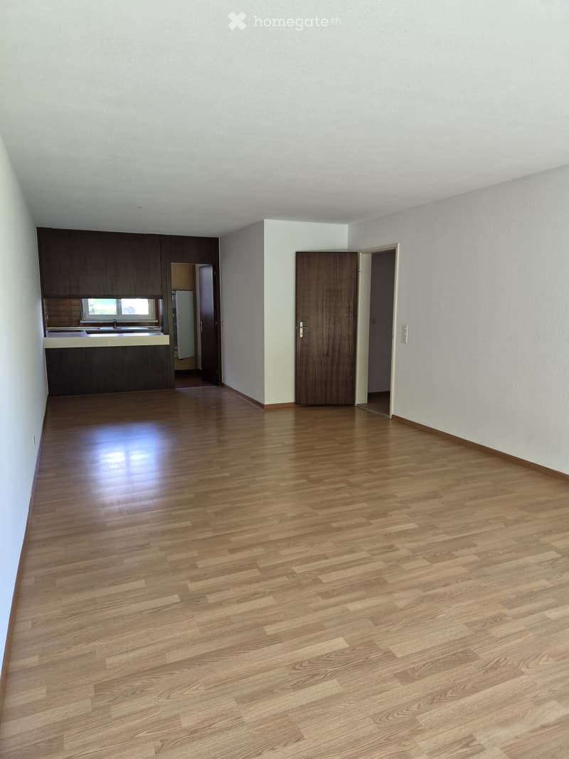 Empty room with wooden flooring, white walls, and a doorway leading to another room. The adjacent room has a kitchen area with cabinets and a sink.