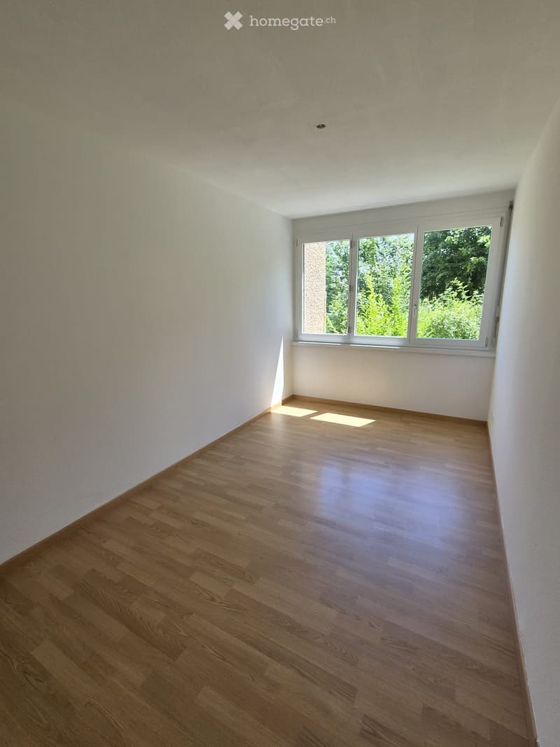 Empty room with wooden flooring and white walls, featuring a large window with a view of greenery outside.