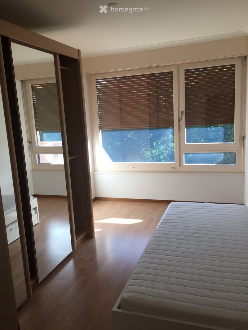 Spacious bedroom with wooden flooring, large windows with blinds, and a neatly made bed with a mattress on a base.